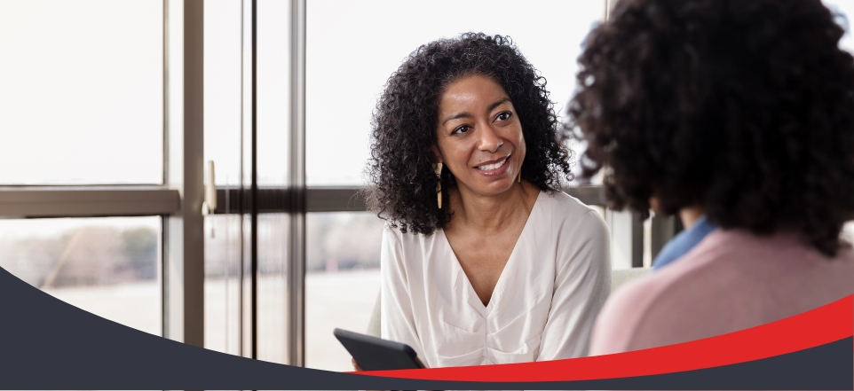Smiling healthcare provider holding a tablet and speaking with a patient in a bright clinic setting, with a red and dark gray curved banner overlay at the bottom.
